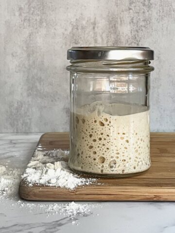 Sourdough starter in a glass jar, fed to its peak, on a cutting board with some flour.