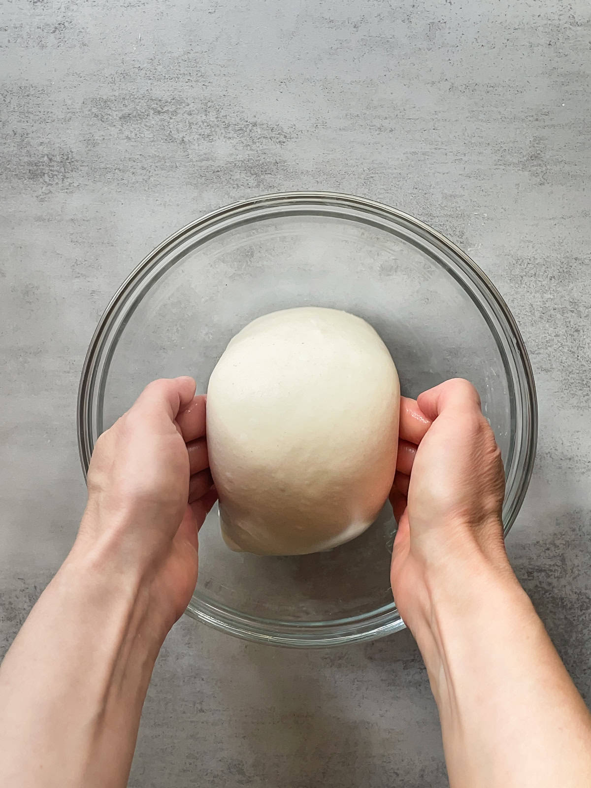 Bread dough lifted with both hands over a glass mixing bowl.