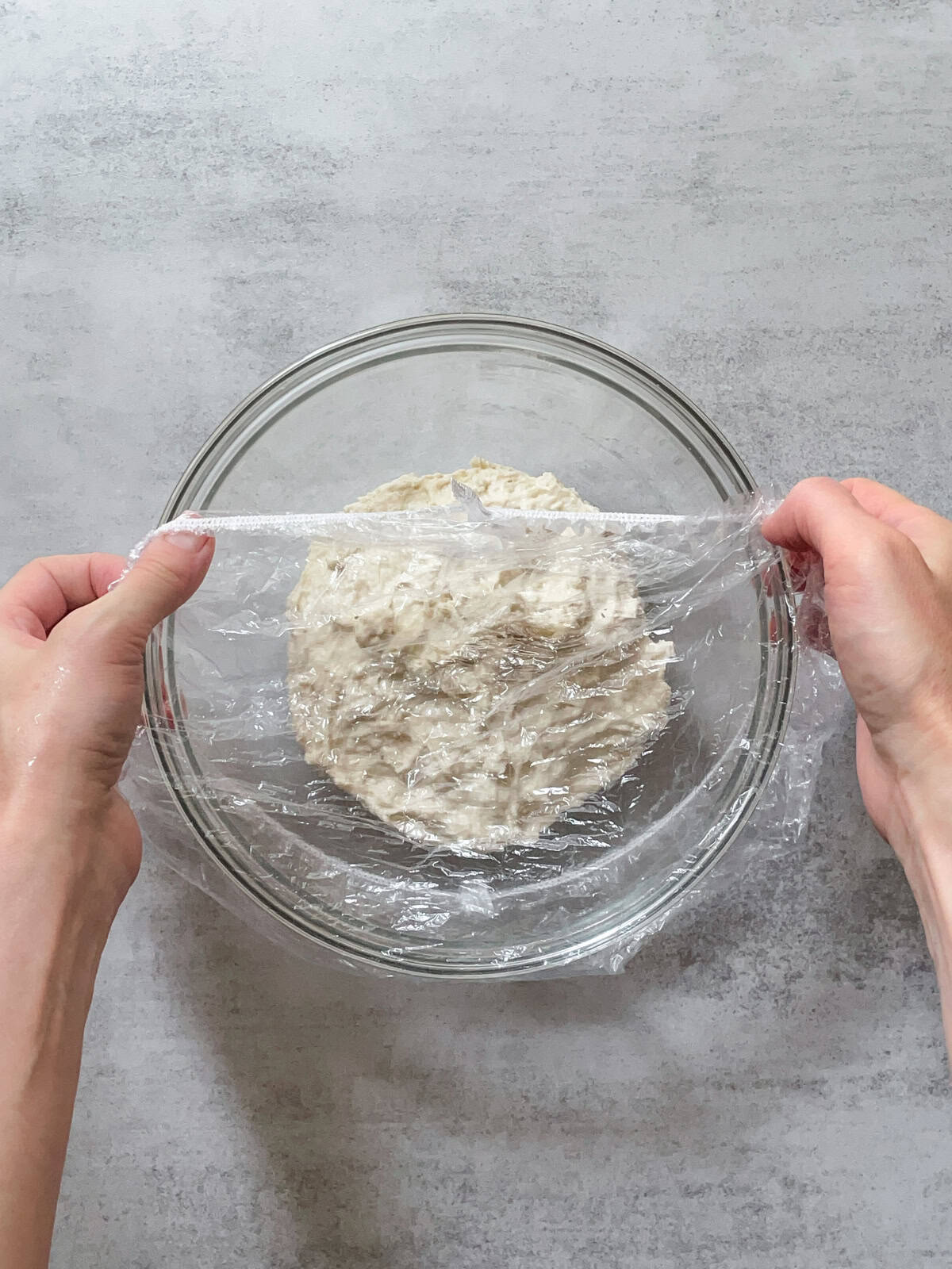 Hands covering a glass bowl of flour and water with plastic cap for autolyse.