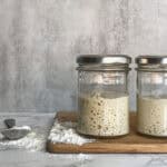 Two jars of sourdough starter with a spoonful of flour on a wooden cutting board.