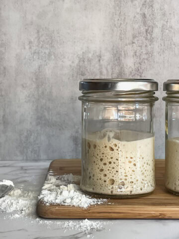 Two jars of sourdough starter with a spoonful of flour on a wooden cutting board.