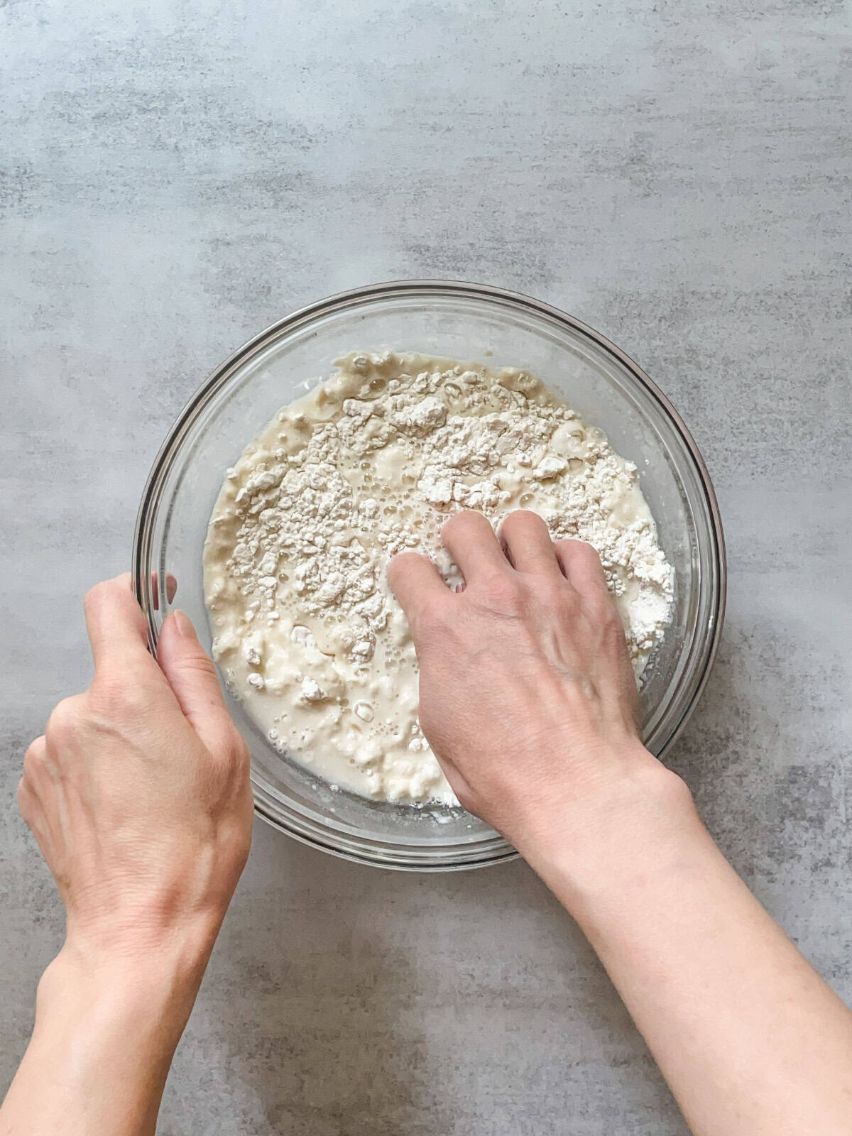 Hands mixing flour and water in a glass bowl for sourdough autolyse.