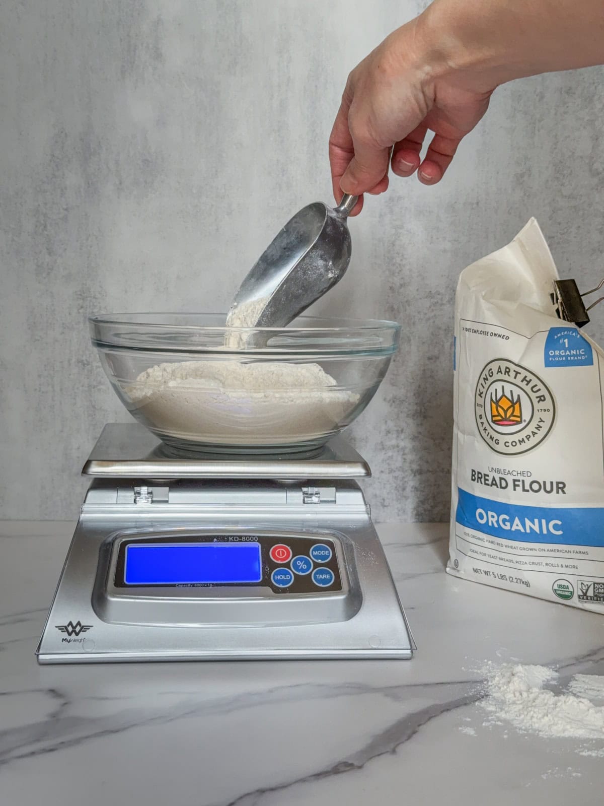 Hand pouring flour into a glass bowl on a kitchen scale.
