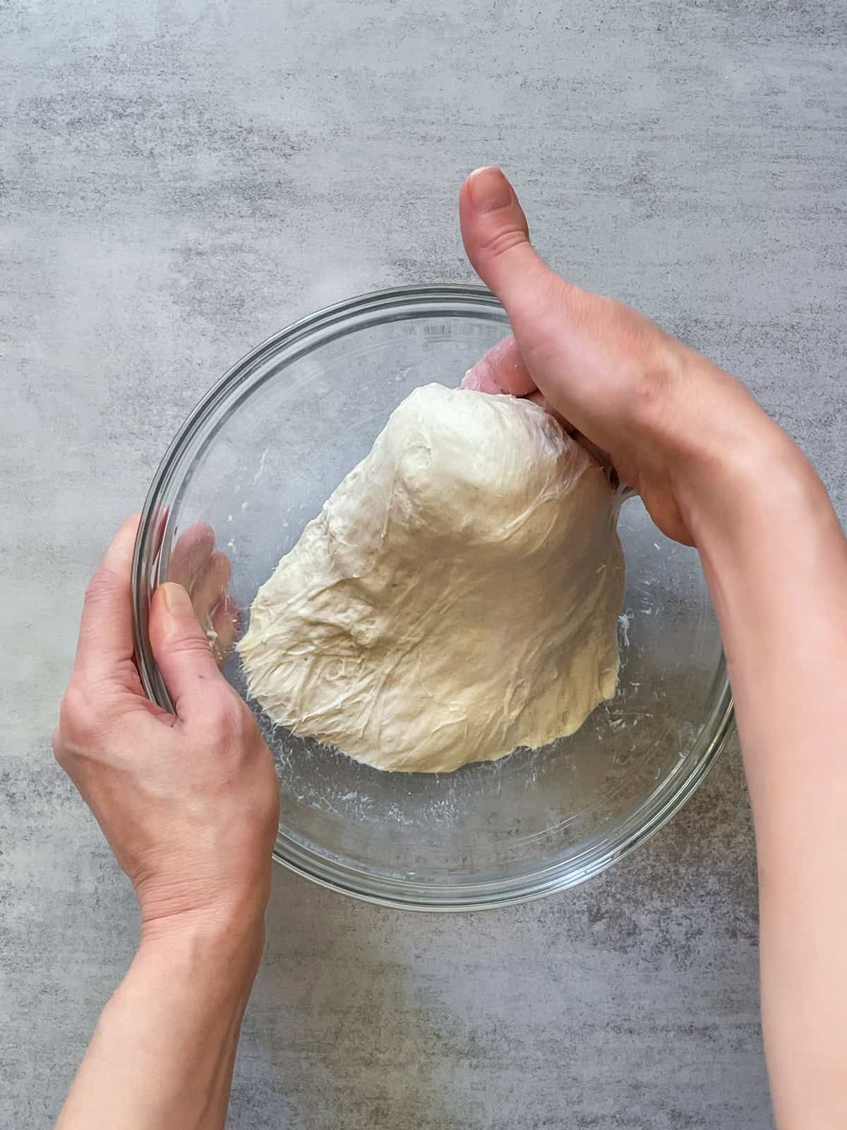A hand lifting bread dough up and to the side in a glass bowl.