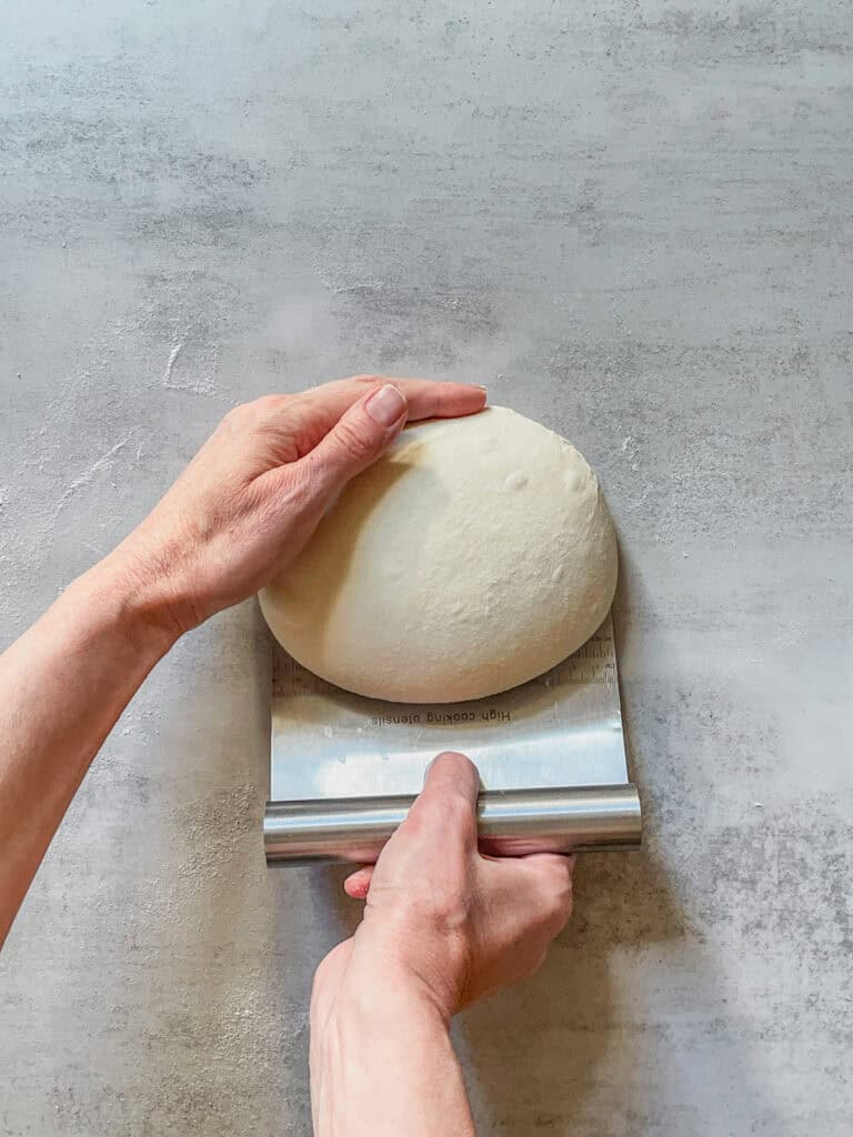 Hands holding a ball of bread dough while using a bench scraper.