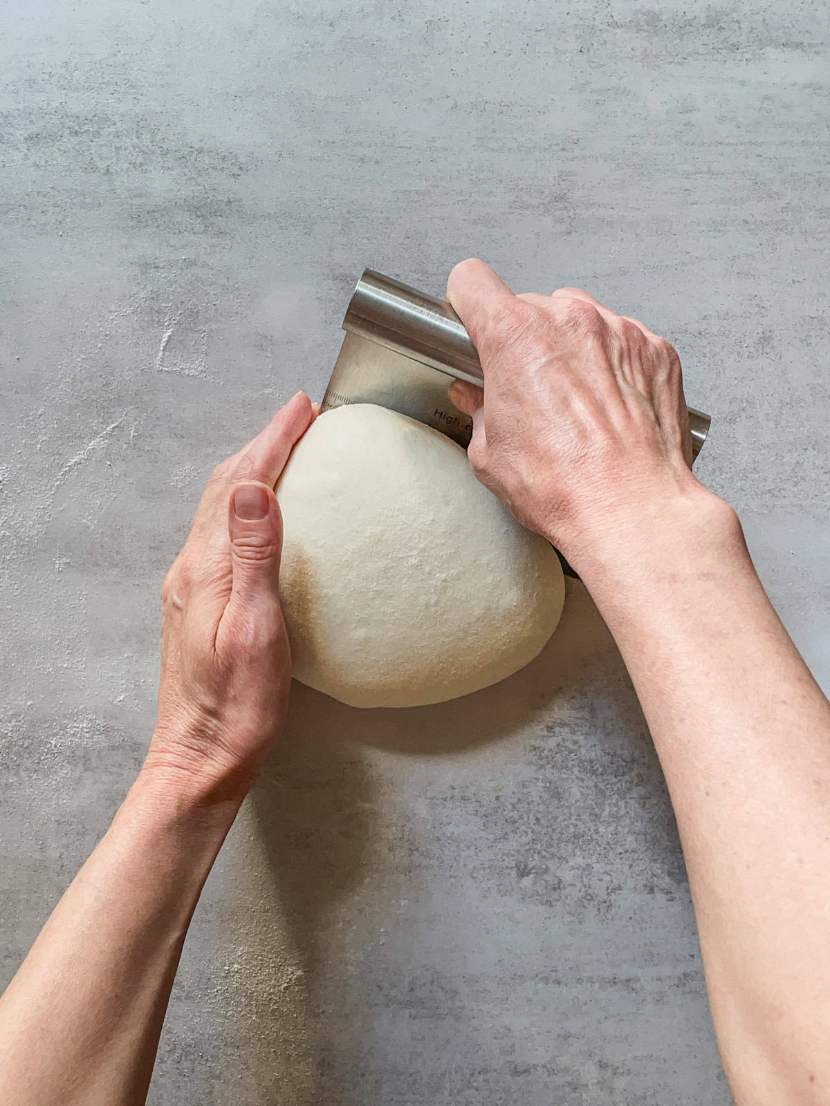 Hands turning a ball of bread dough while using a bench scraper.