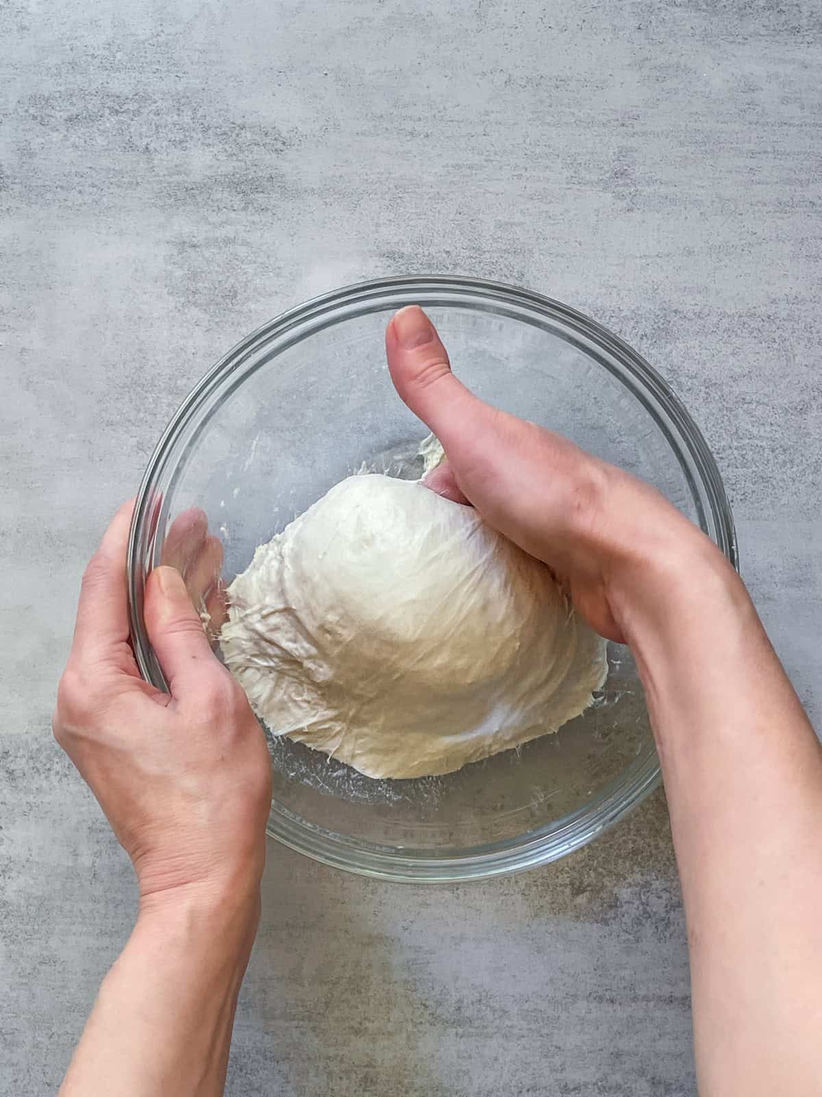 Hand lifting bread dough in a glass bowl.