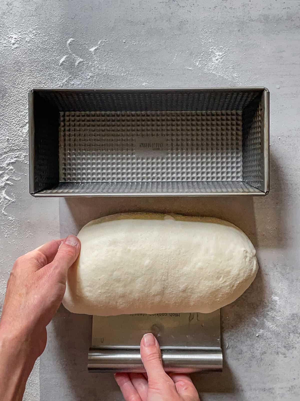 Hands using a bench scraper to flip the batard into a greased loaf pan.