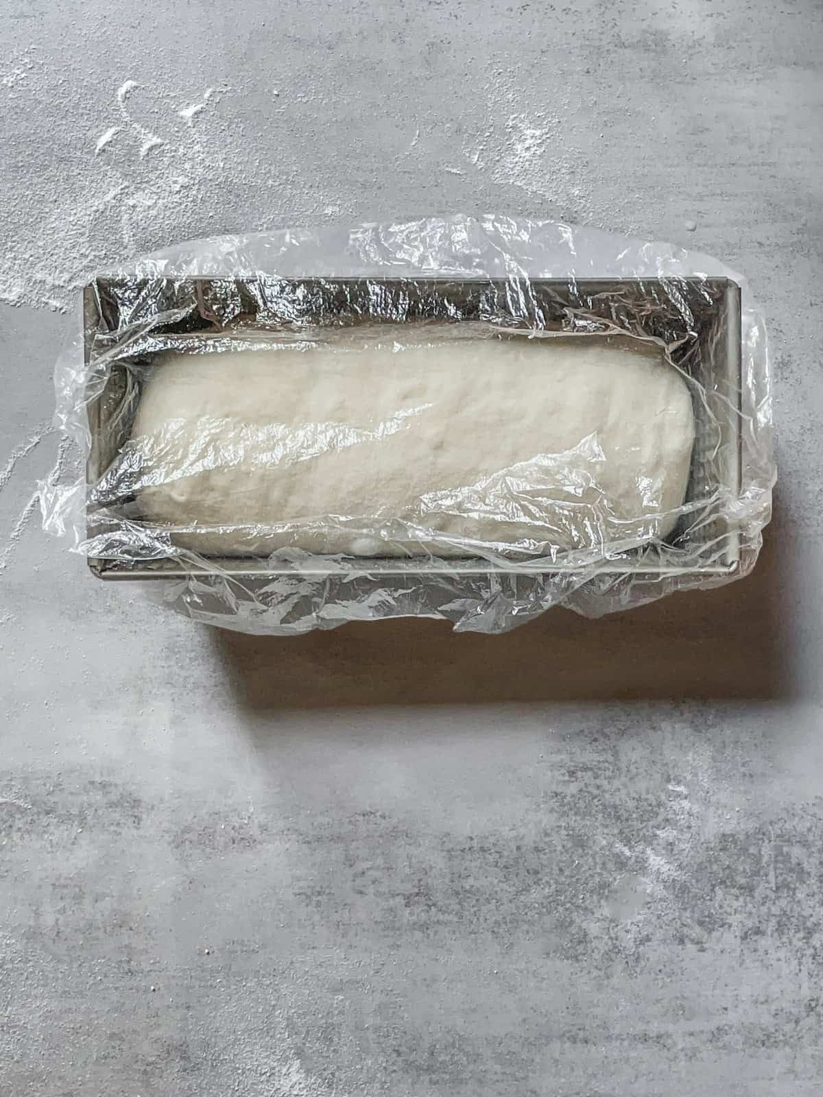 Loaf pan with shaped dough covered with plastic.