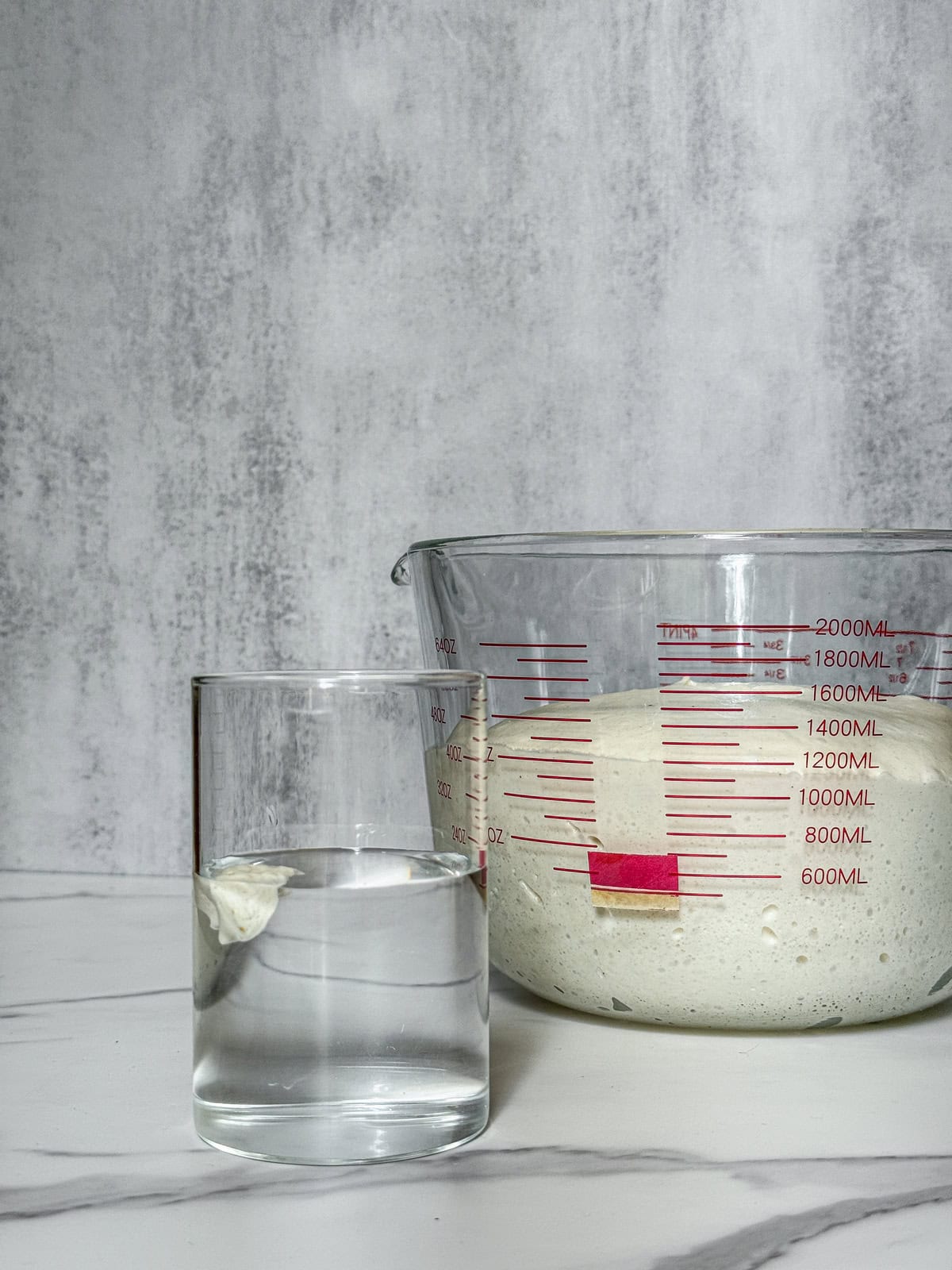 Dough floating in a glass of water next to a larger measuring cup, front view.