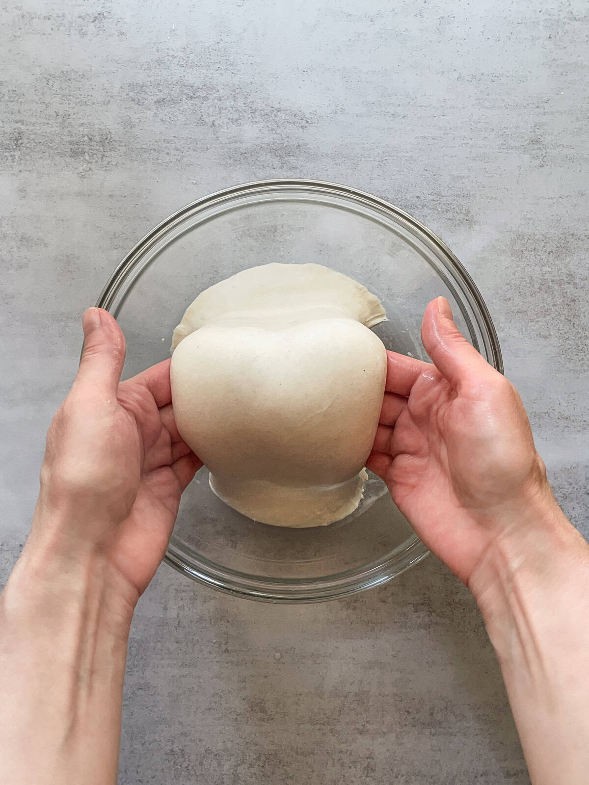 Hands lifting dough over a glass mixing bowl.
