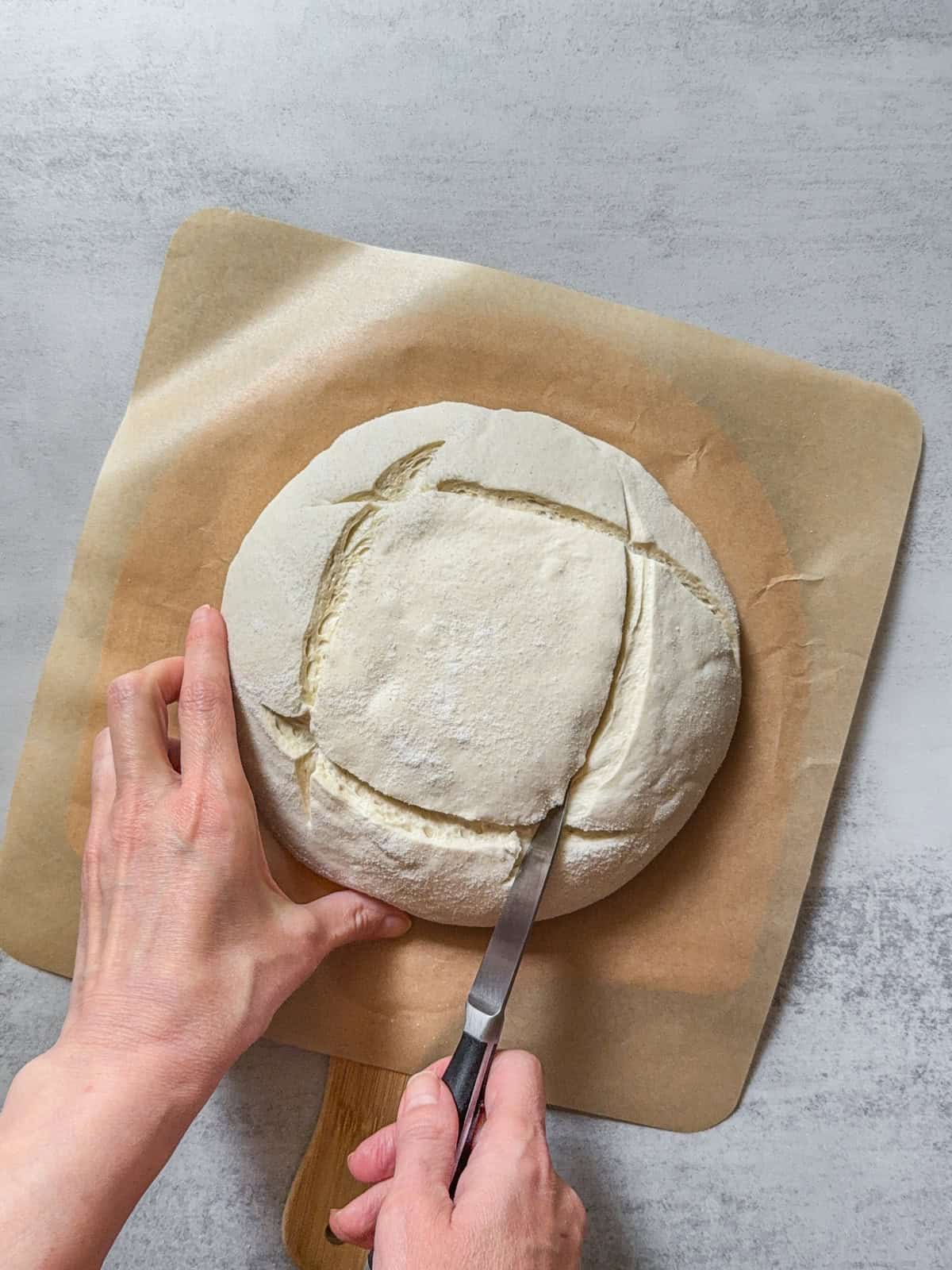 Hands holding a dough boule while scoring it with a knife.