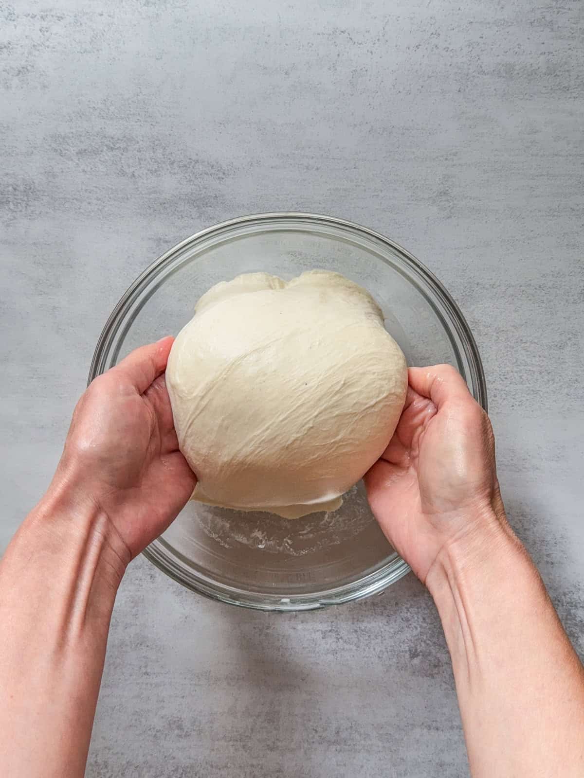 Hands lifting bread dough over a glass mixing bowl.
