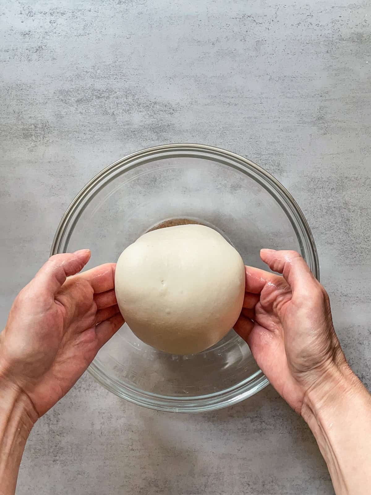 Hands lifting bread dough in a glass bowl.