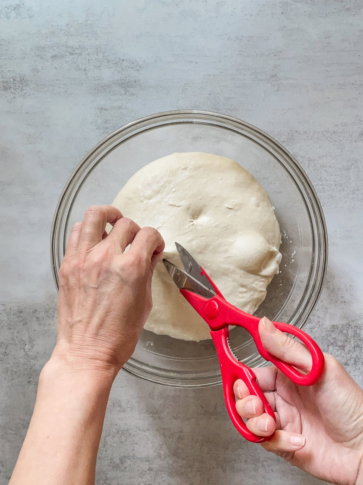 Hands snipping a piece of dough with scissors over a glass bowl.
