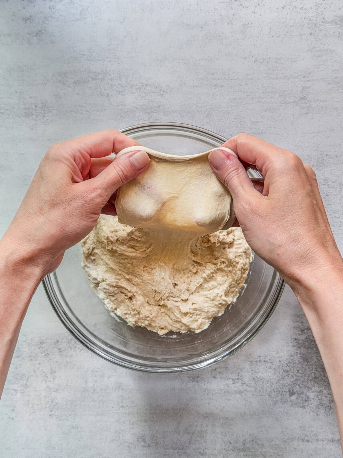 Hands stretching sourdough dough over a glass bowl after autolyse.