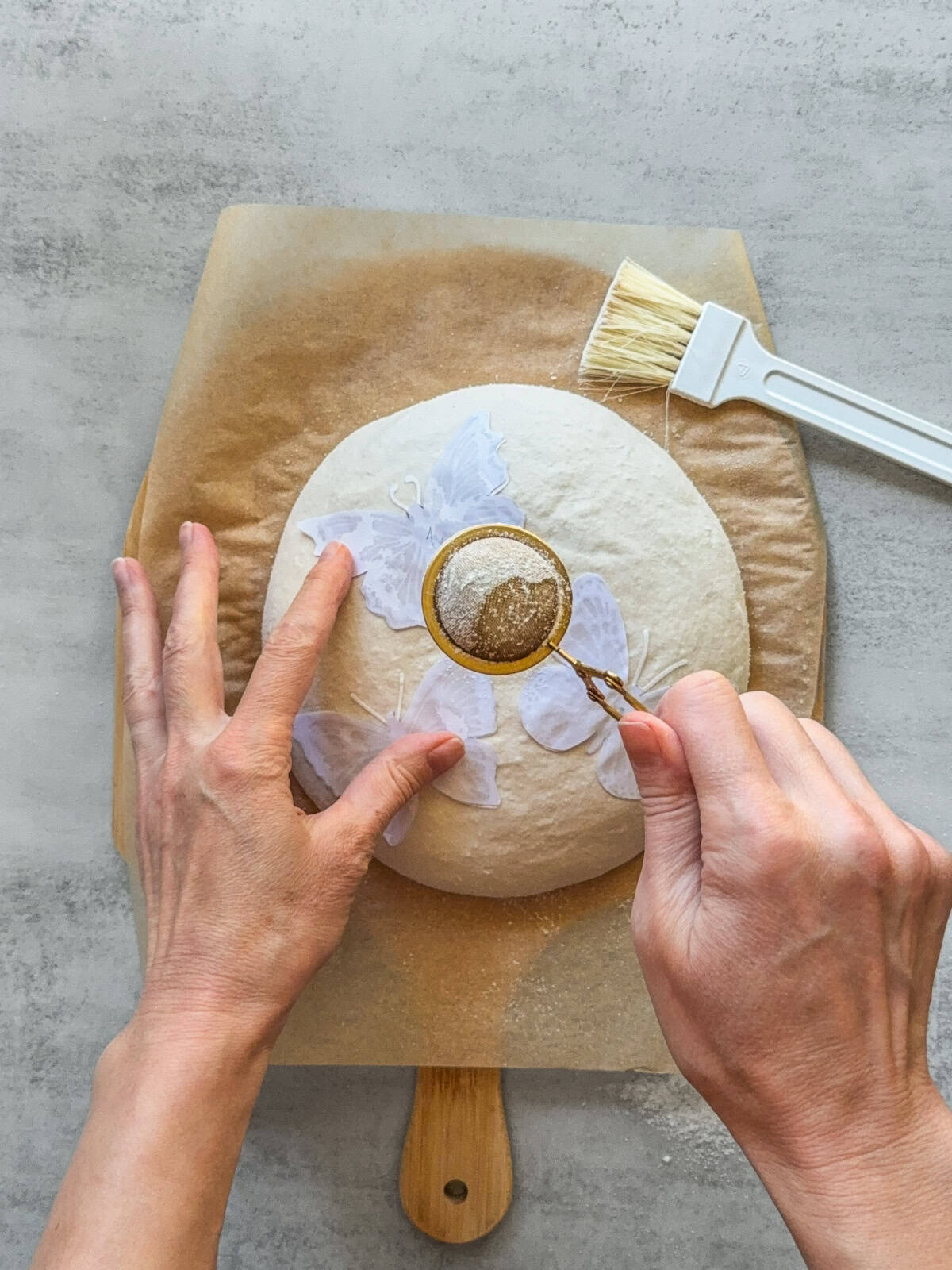 Hands dusting flour over the dough using a flour duster with a stencil in place.