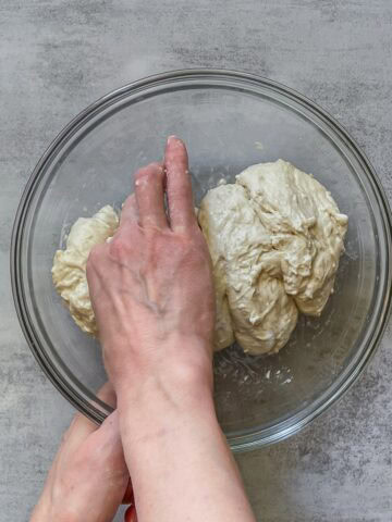 A hand pinching sourdough dough with thumb and forefinger using the pincer method in a glass bowl.